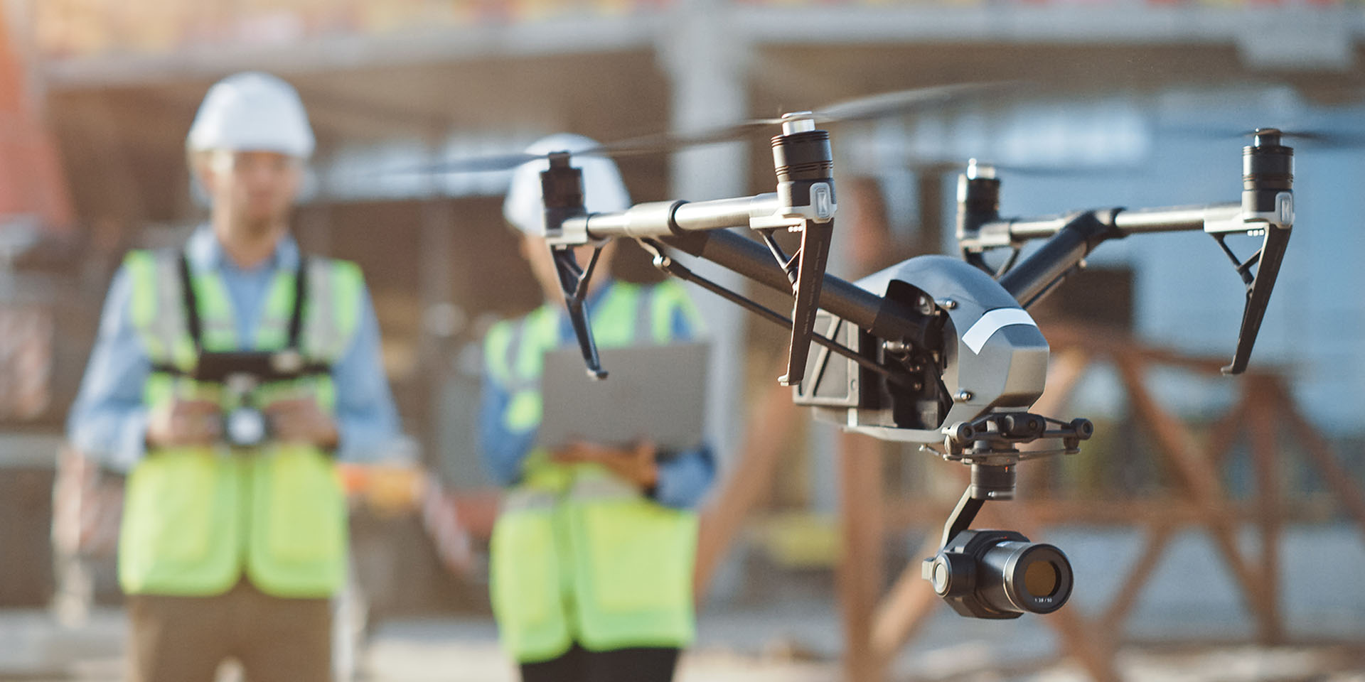Drohne im Einsatz auf einer Baustelle, gesteuert von zwei Mitarbeitenden mit Schutzhelm und Warnweste zur Inspektion.